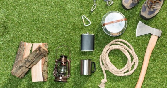 Sprzęt survivalowy ułożony na trawie: siekiera, sznurek, buty trekkingowe, latarnia, drewno opałowe, menażka, karabinki i kubek metalowy.